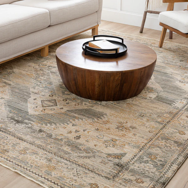 Round wooden coffee table with a dark finish, placed on a patterned area rug featuring muted tones of beige, gray, and light brown. The table has a black tray with a book on top. Surrounding the table are a light beige sofa and a chair with wooden legs and white cushions. The setting is in a room with light wood flooring and white walls.