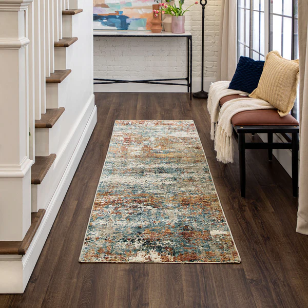A narrow runner rug with a distressed, multicolored pattern featuring shades of blue, orange, beige, and brown lies on a dark wood floor in a hallway. To the right, a bench with a brown leather cushion holds a cream throw blanket and two textured pillows, one navy blue and one mustard yellow. A console table with a black metal frame and wooden top is against a white brick wall at the end of the hallway, decorated with a colorful abstract painting and a vase with flowers. A staircase with white risers and wooden treads is on the left side.