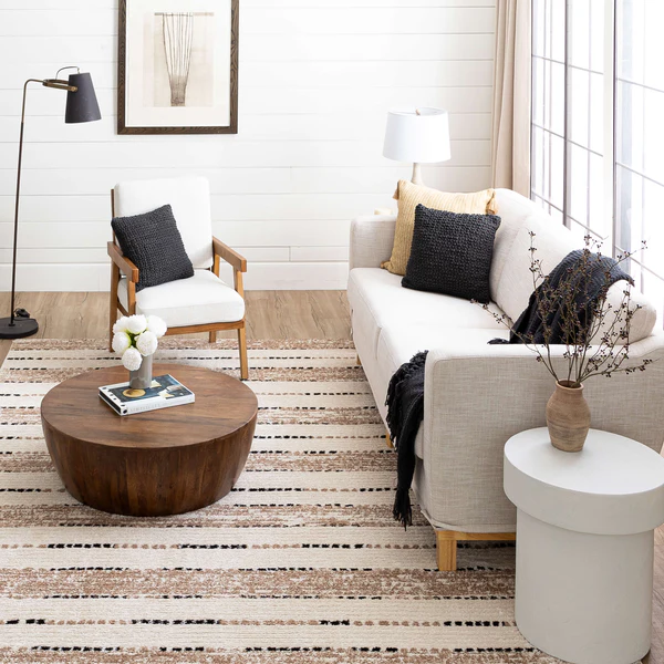 A modern living room with a light beige sofa adorned with black and beige cushions and a dark throw. Next to the sofa is a white round side table with a beige vase holding dried branches. A wooden armchair with white cushions and a black pillow sits beside a round wooden coffee table topped with a small vase of white flowers and a book. The floor features a striped beige and brown rug, and the walls are white with horizontal paneling. Two floor lamps, one black and one white, provide lighting, and a framed abstract artwork hangs on the wall. Large windows with beige curtains allow natural light into the room.