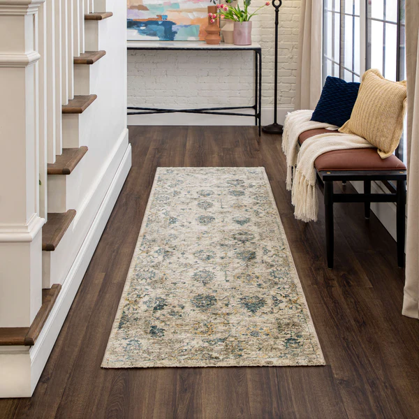 A narrow runner rug with a distressed vintage pattern in muted tones of beige, blue, and gray lies on dark wood flooring. To the right, a bench with a brown leather cushion is adorned with a cream throw and two textured pillows, one navy blue and one mustard yellow. On the far side, a slim black metal console table holds a pink vase with flowers and a colorful abstract painting hangs above it. A staircase with white risers and wooden treads is on the left.
