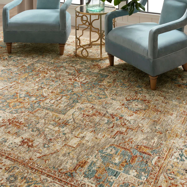 A patterned area rug with a distressed, vintage look featuring muted tones of beige, blue, rust, and brown. Two light blue upholstered armchairs with wooden legs are positioned on the rug, along with a small round gold metal side table with a glass top. The setting is near a window with natural light.