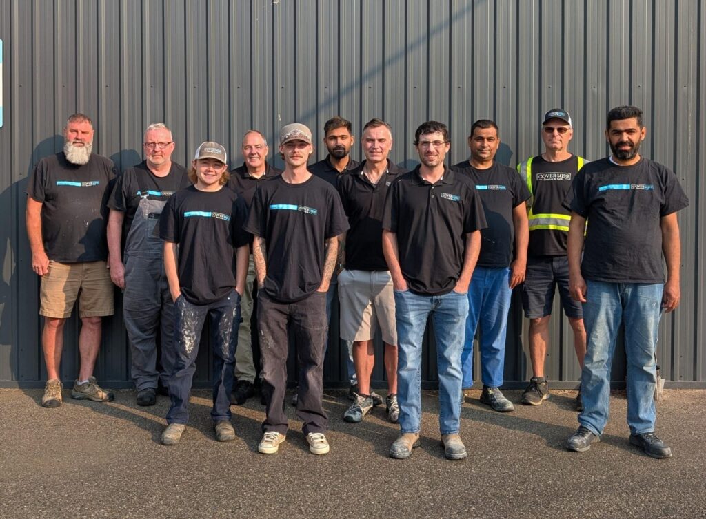 Nine men in black company shirts stand in a row in front of a gray corrugated metal wall.